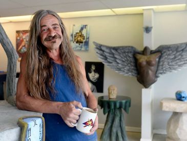 A man with long hair holding a coffee mug in an art studio.
