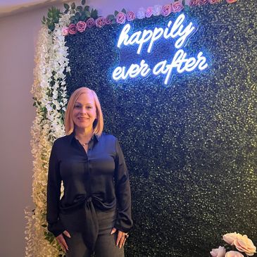 Woman posing in front of a floral wall with "happily ever after" sign.