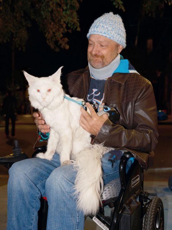 Man in a wheelchair holding a large white cat at night.