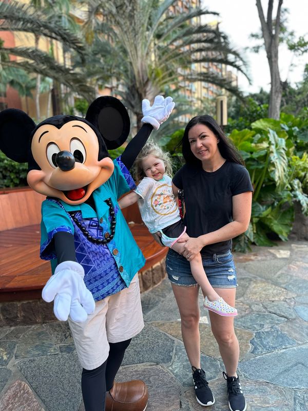A joyful child with her mother posing with Mickey Mouse in a tropical outdoor setting.