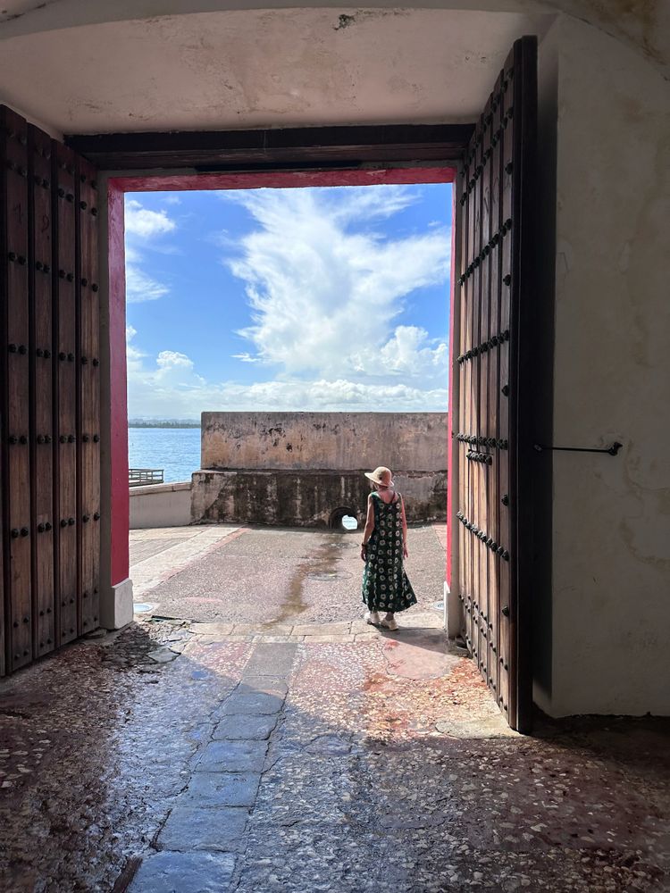 A woman in a floral dress and hat walking outside through a large open doorway.