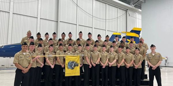 A group of uniformed cadets posing in front of jets inside a hangar.