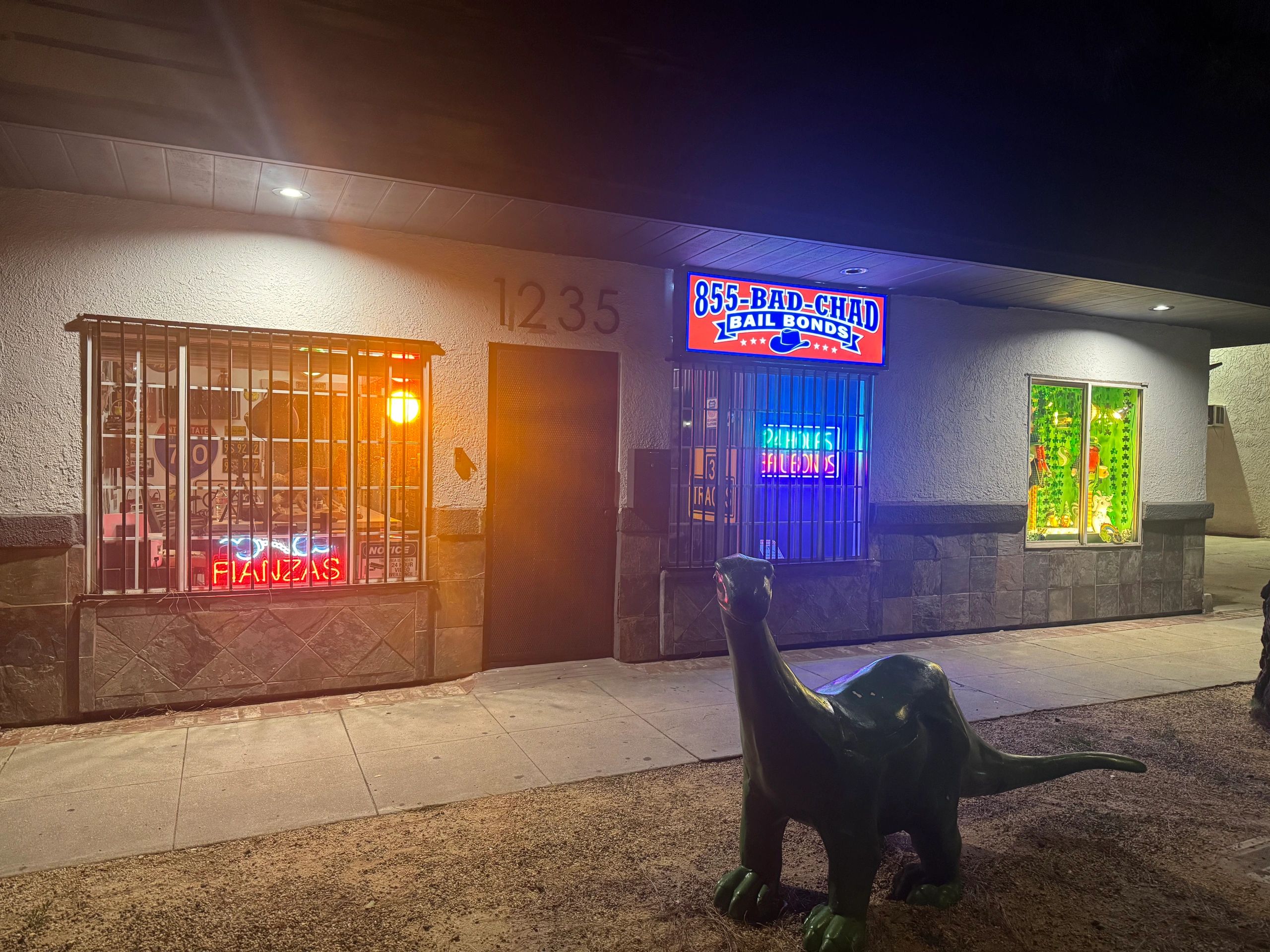 Night view of a bail bonds office with a dinosaur statue outside.