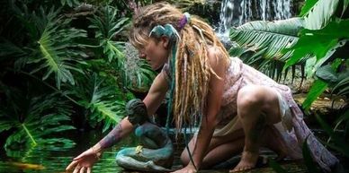 A woman with dreadlocks reaches out to touch water in a lush jungle setting.