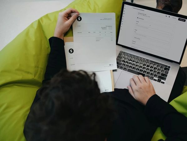 Person reviewing documents and working on a laptop, possibly handling invoices or reports.