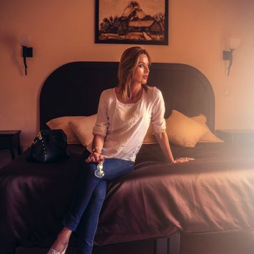 A woman sits on a bed holding a wine glass, looking thoughtfully to the side.