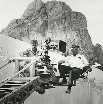 Two men operating a film camera on a crane with a mountain backdrop.