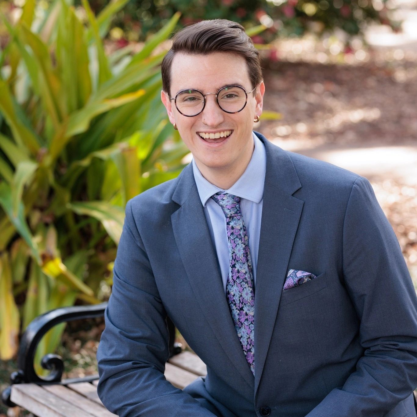 Smiling young man in a blue suit sitting on a bench outdoors.
