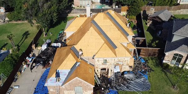 Workers replacing the roof on a large suburban house under clear skies.