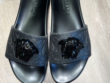 Black Versace slides with Medusa emblem on a wooden floor.