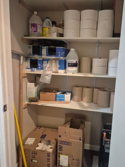 Storage closet with shelves holding toilet paper, cleaning supplies, and cardboard boxes.