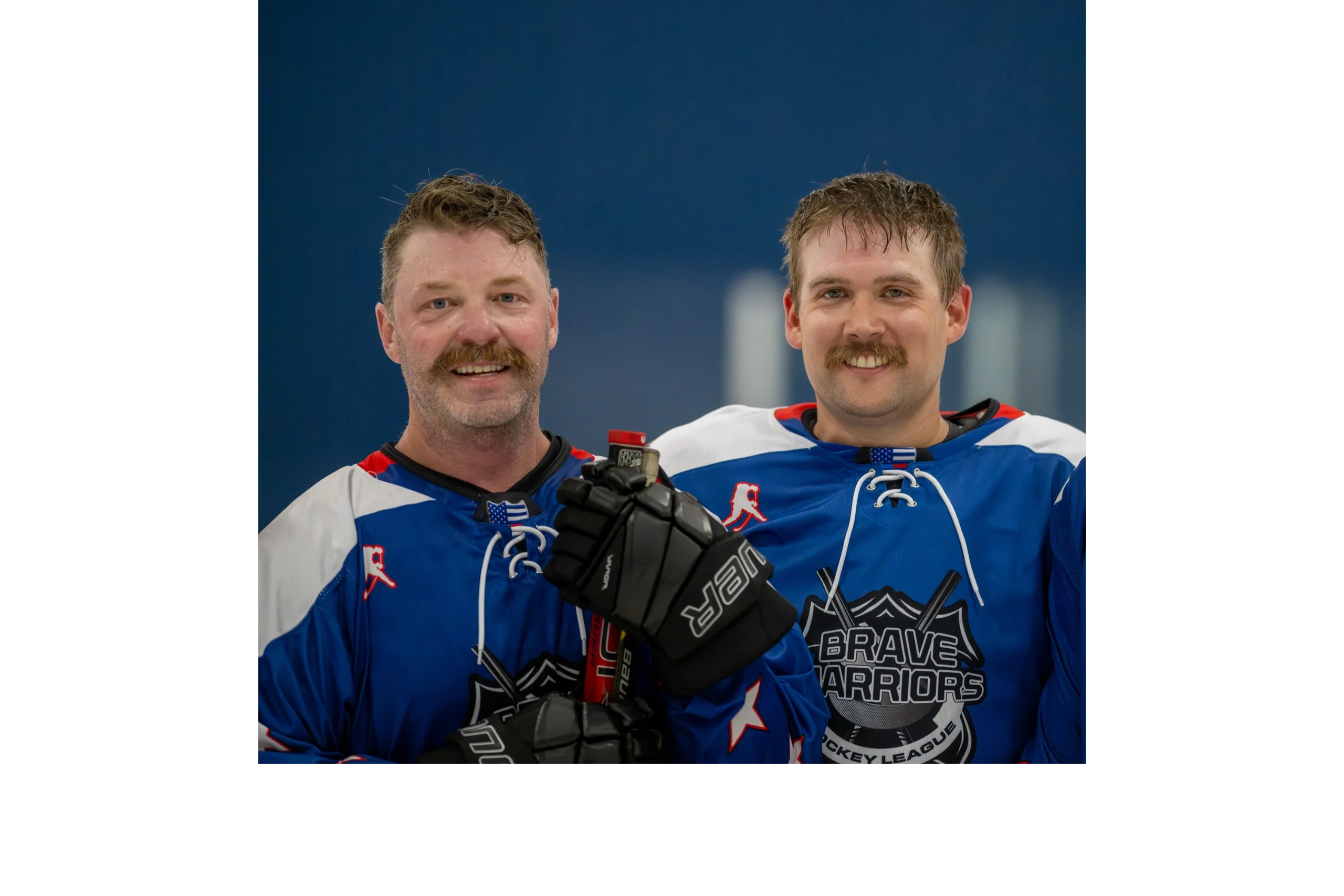 Two smiling first responder and veteran hockey players in blue jerseys with mustaches pose together 