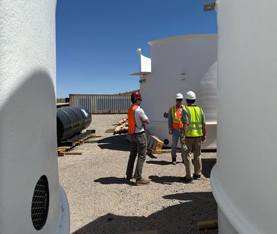 Three construction workers in safety gear discussing at an industrial site with large white tanks.