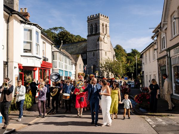 Wedding parade with band and guests on village street.
