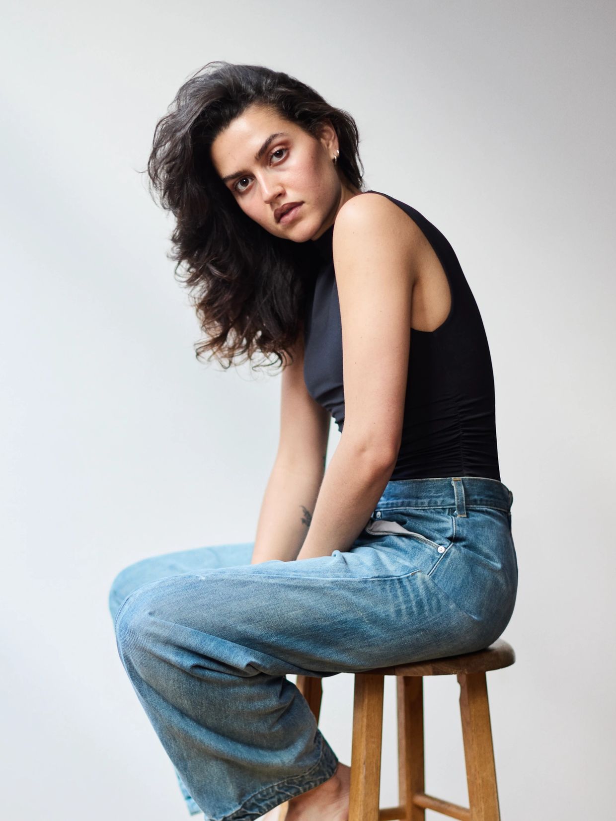 Woman with dark hair sitting on a wooden stool in casual attire.