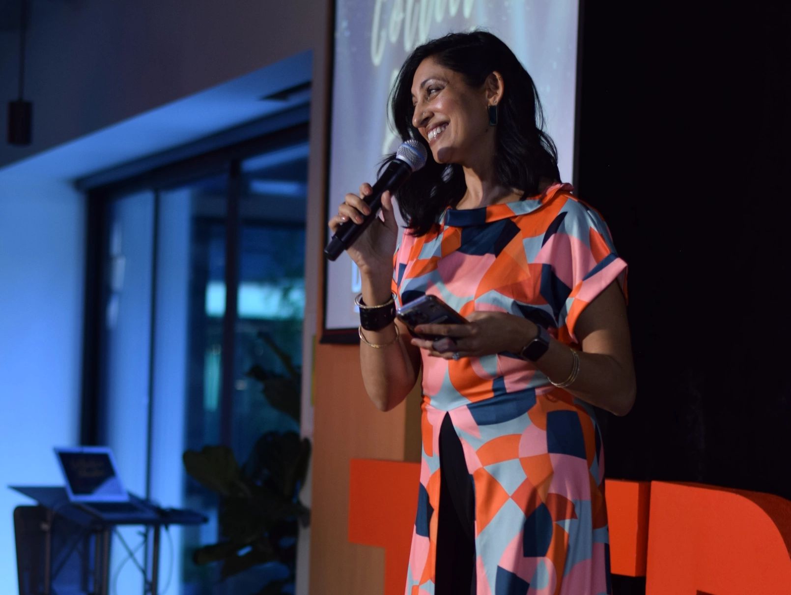 Woman speaking on stage with a microphone, smiling warmly.