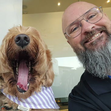 Man with glasses and beard smiles next to a yawning dog wearing a striped bandana.