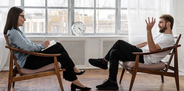 <img src="therapist-and-patient-session" alt="Woman sitting opposite man in two wooden chairs">