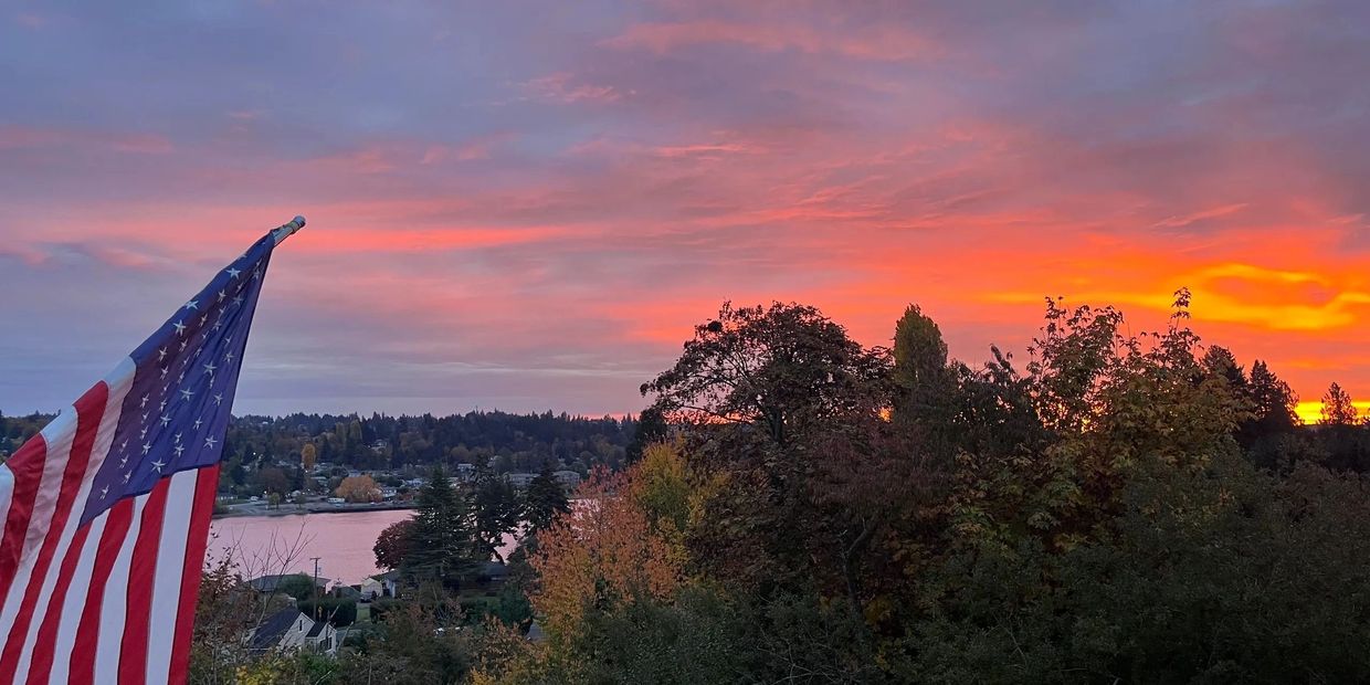 American flag flying with a vibrant sunset over trees and water.