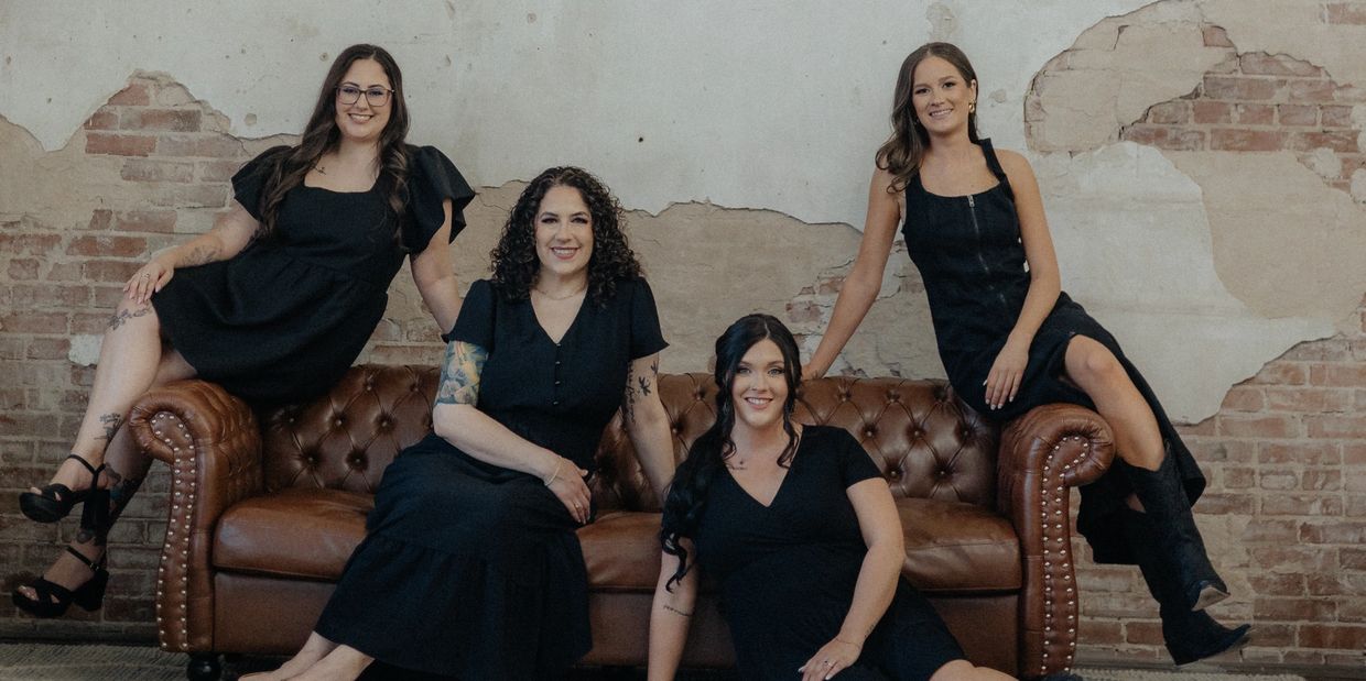 Four women in black dresses posing on and around a brown leather couch against a rustic brick wall.