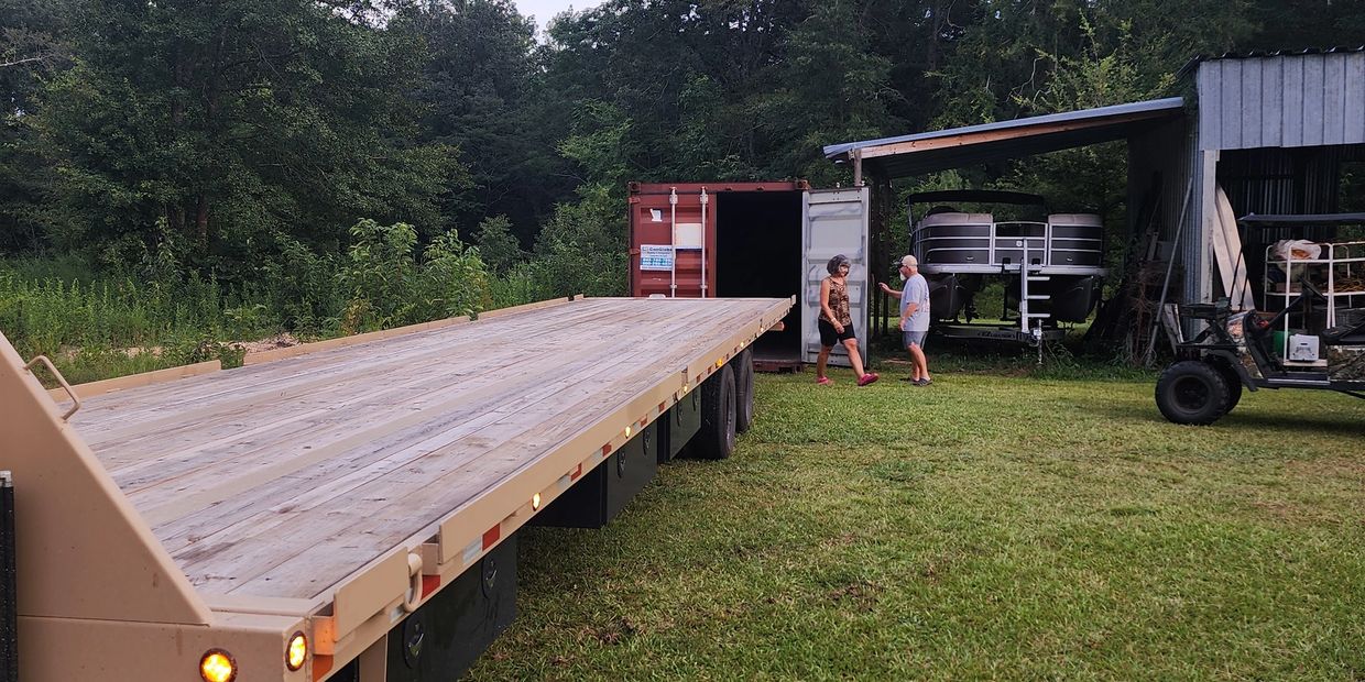 Two people standing near a flatbed trailer and a boat under a shed in a grassy area.