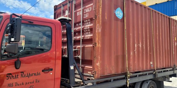 Red truck carrying a large rusty shipping container under a blue sky.