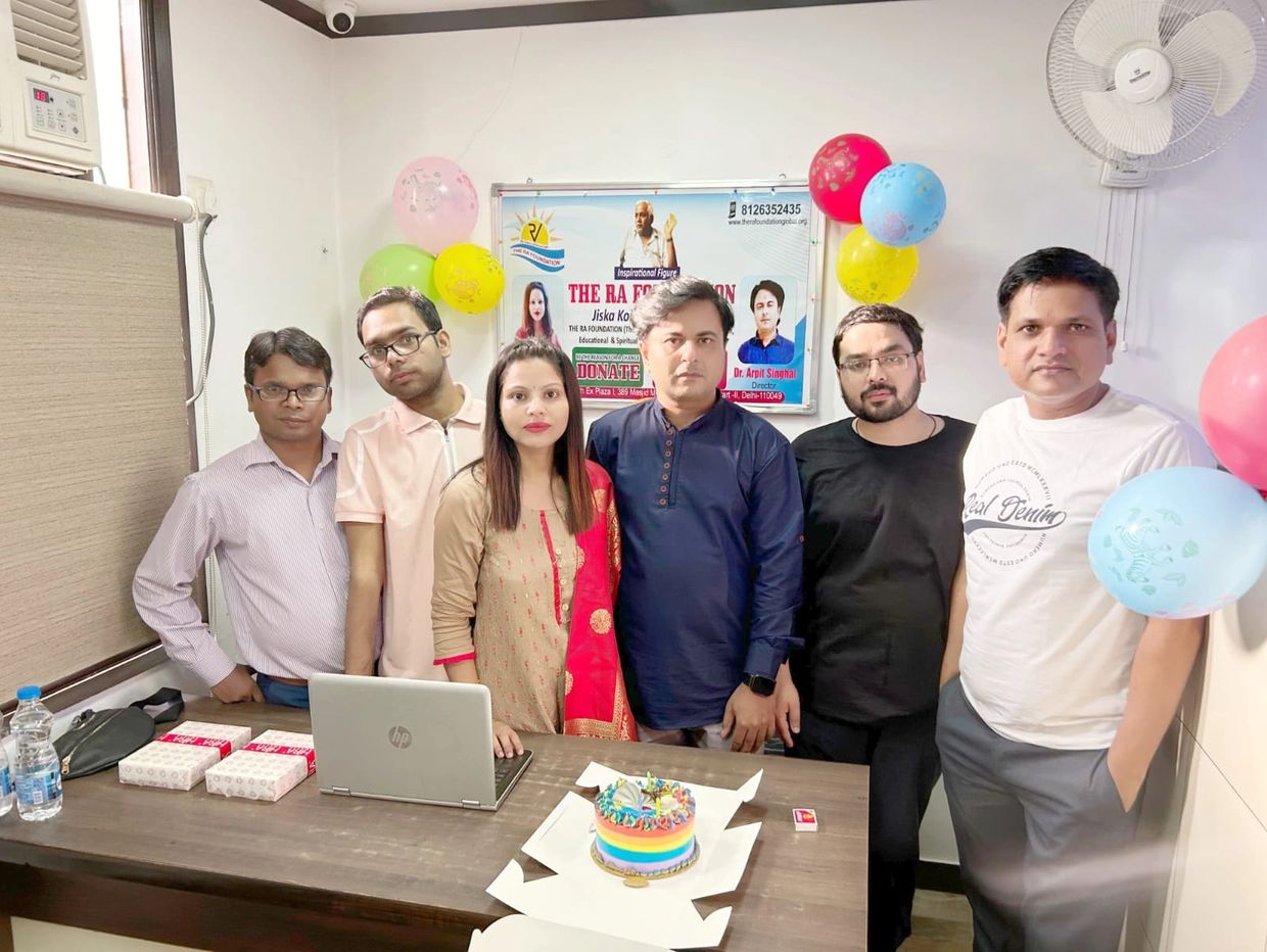 Six people standing behind a table with a laptop and a rainbow cake, decorated with colorful balloons.