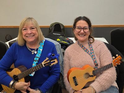 Two smiling women holding ukuleles, sitting side by side indoors.