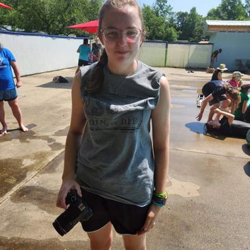 A young woman holding a camera at an outdoor event on a sunny day.