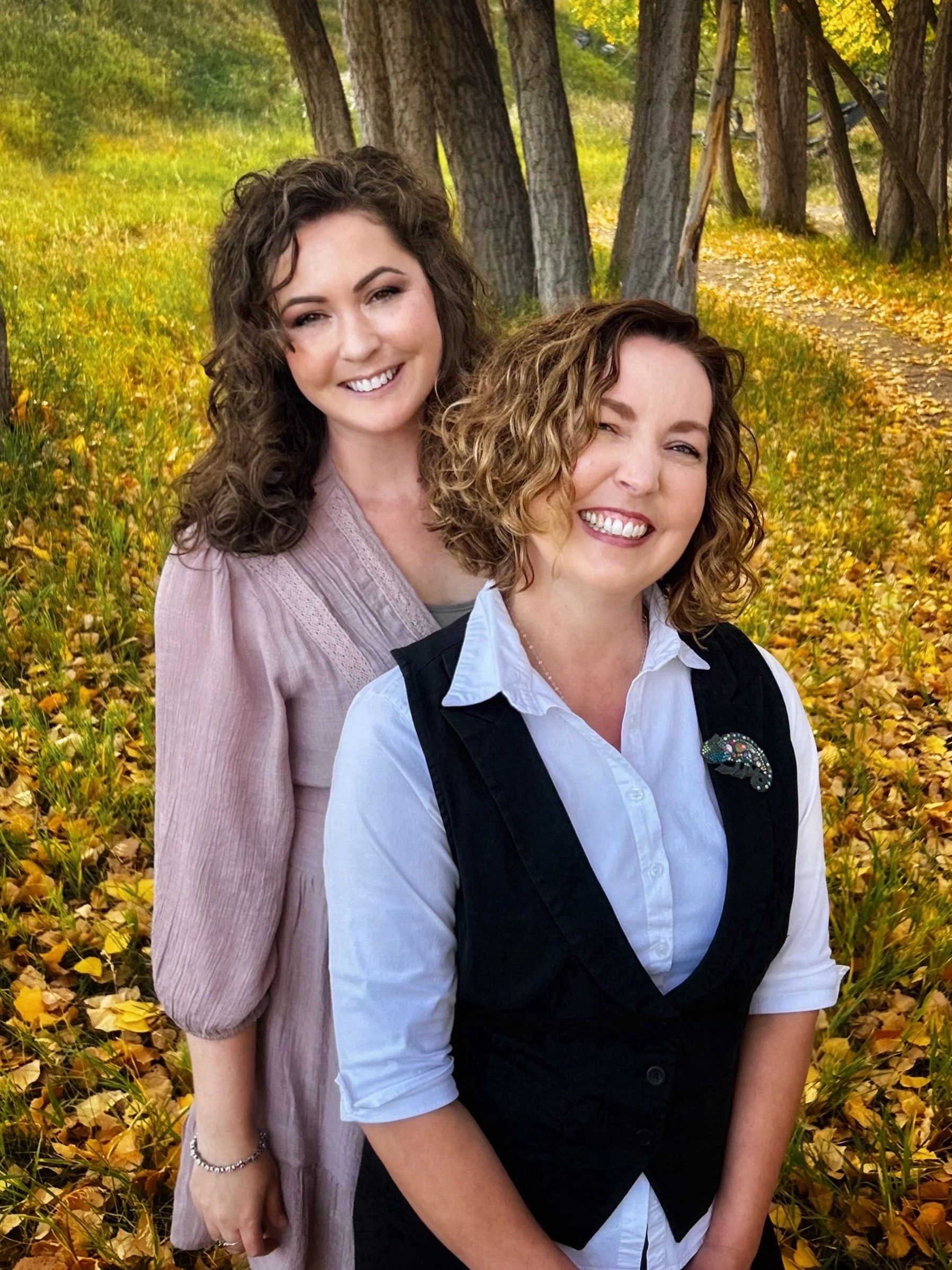 Two women smiling outdoors in a fall setting with yellow leaves and trees.