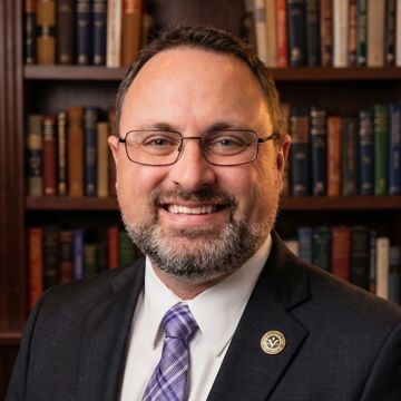 Man in suit and glasses smiling in front of a bookshelf.