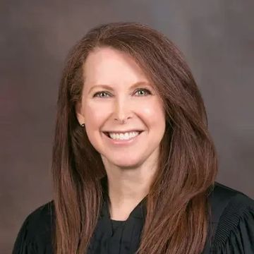 Smiling woman with long brown hair in black robe against a neutral background.