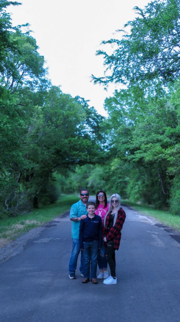 A happy family of four stands on a tree-lined road, smiling at the camera.