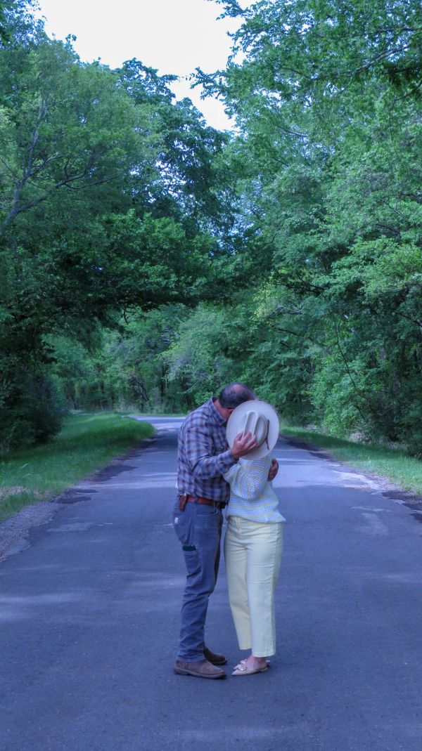 Couple sharing a kiss on a tree-lined road, hiding behind a hat.