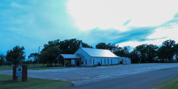 A small white church with a metal roof and empty parking lot under a cloudy sky.