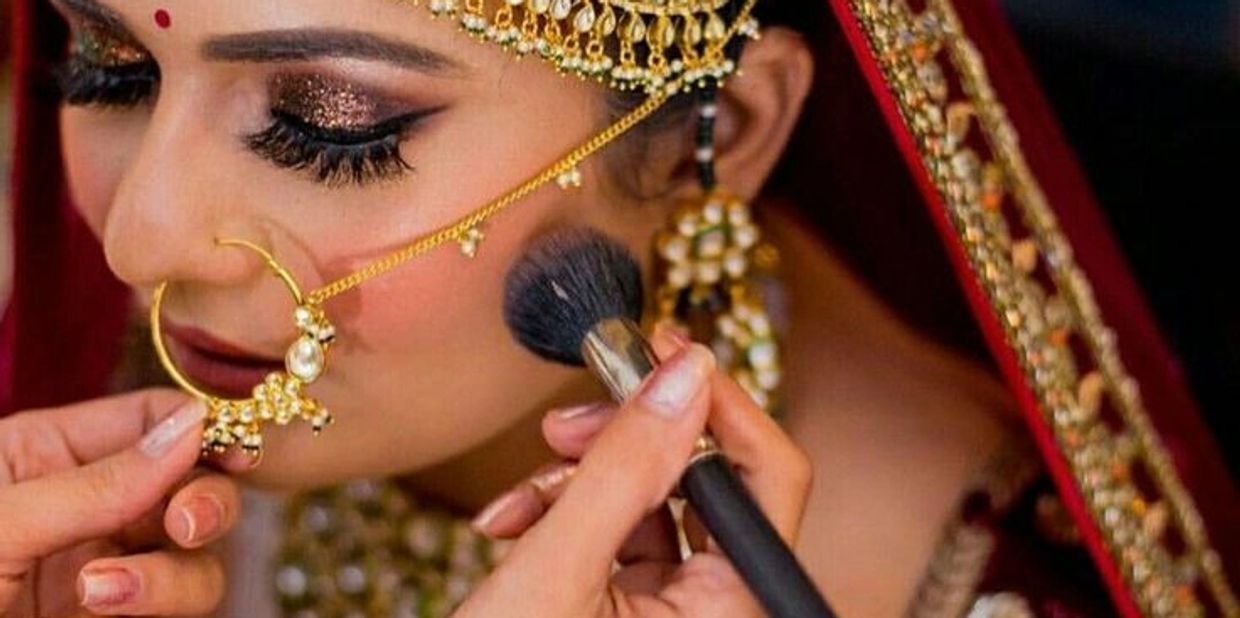 Bride getting makeup applied, wearing traditional Indian jewelry and attire.