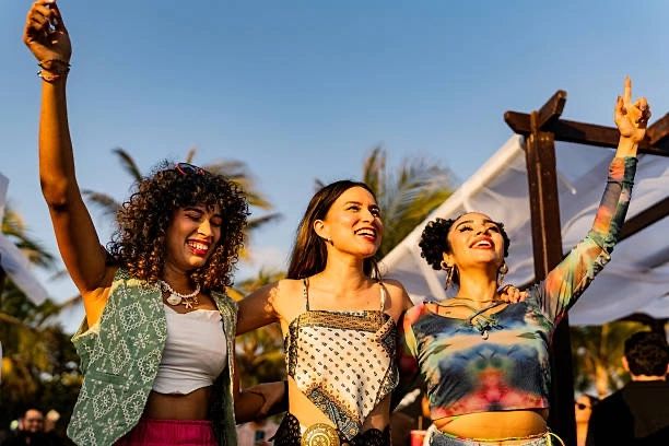 Three women joyfully dancing outdoors in vibrant clothing under blue sky.
