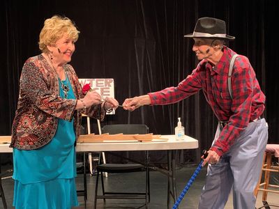 Two elderly actors performing a scene on stage with props.