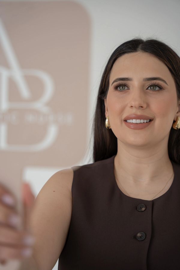 Smiling woman in a dark sleeveless top holding a white object, with a blurred logo background.