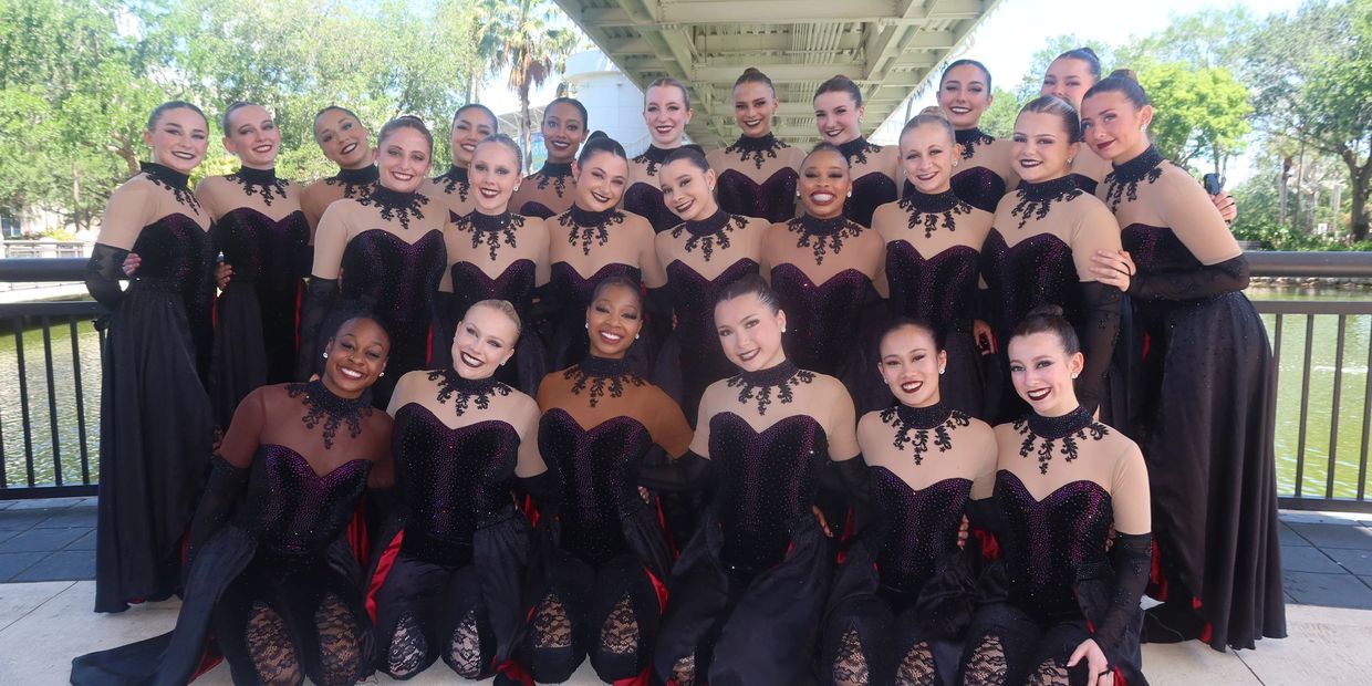 A large group of dancers in matching elegant black and nude costumes posing outdoors by the water.