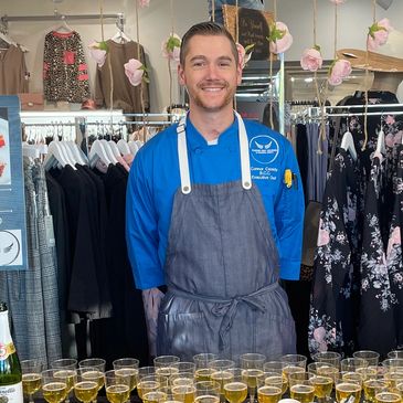 Smiling chef in blue and gray apron stands behind table with drinks in a clothing store.