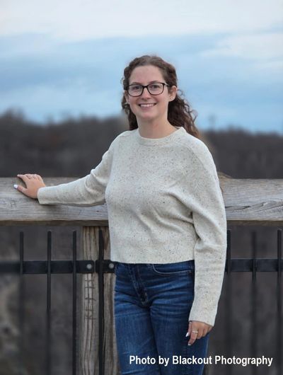 Young woman in glasses and sweater smiling outdoors by a wooden fence.