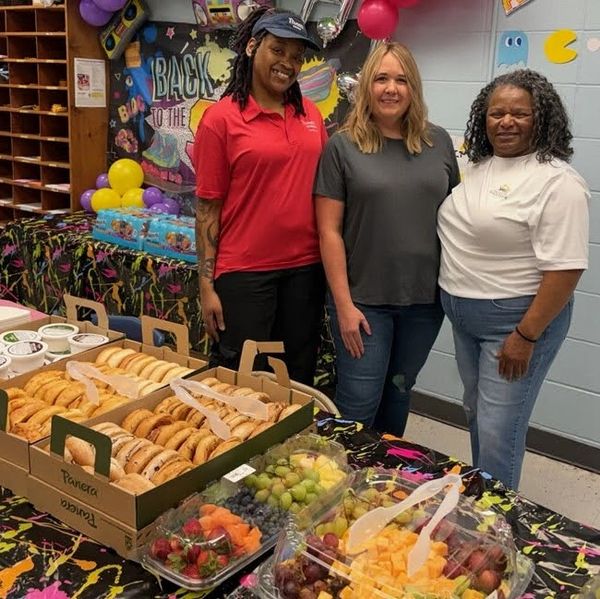 Three women smiling beside a table with bagels and fruit trays.