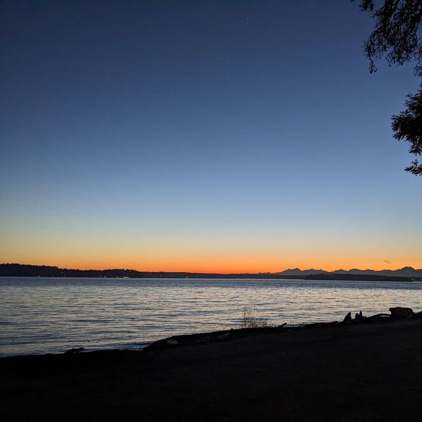 Silhouetted figures by the water at sunset with mountains in the distance.