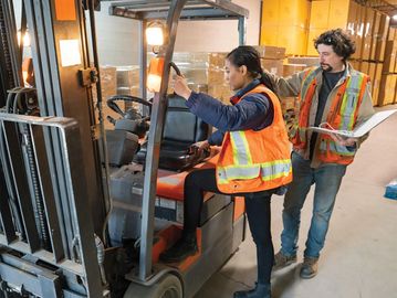 Two workers operating a forklift in a warehouse with safety vests.