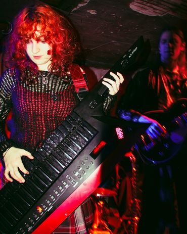 Red-haired musician playing a keytar on stage with a guitarist in the background.