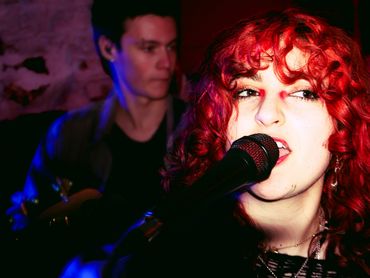 A red-haired woman singing into a microphone with a guitarist in the background.