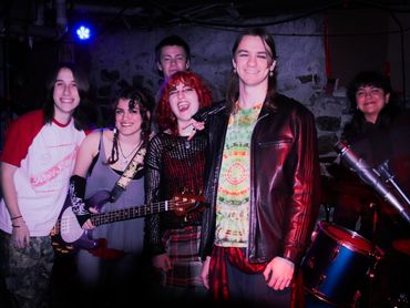 A group of six young musicians posing together in a dimly lit room with musical instruments.