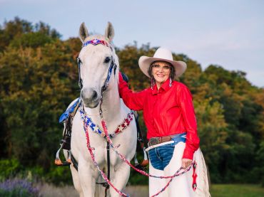 A woman in a red shirt and white cowboy hat stands beside a white horse.