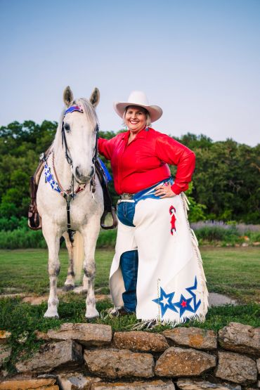 Woman in cowboy attire stands confidently with a white horse outdoors.